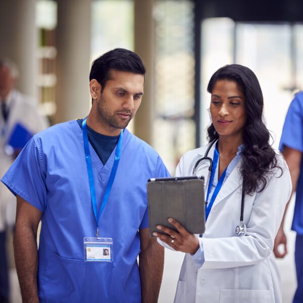 Two Medical Staff In White Coats And Scrubs With Digital Tablet Having Informal Meeting In Hospital